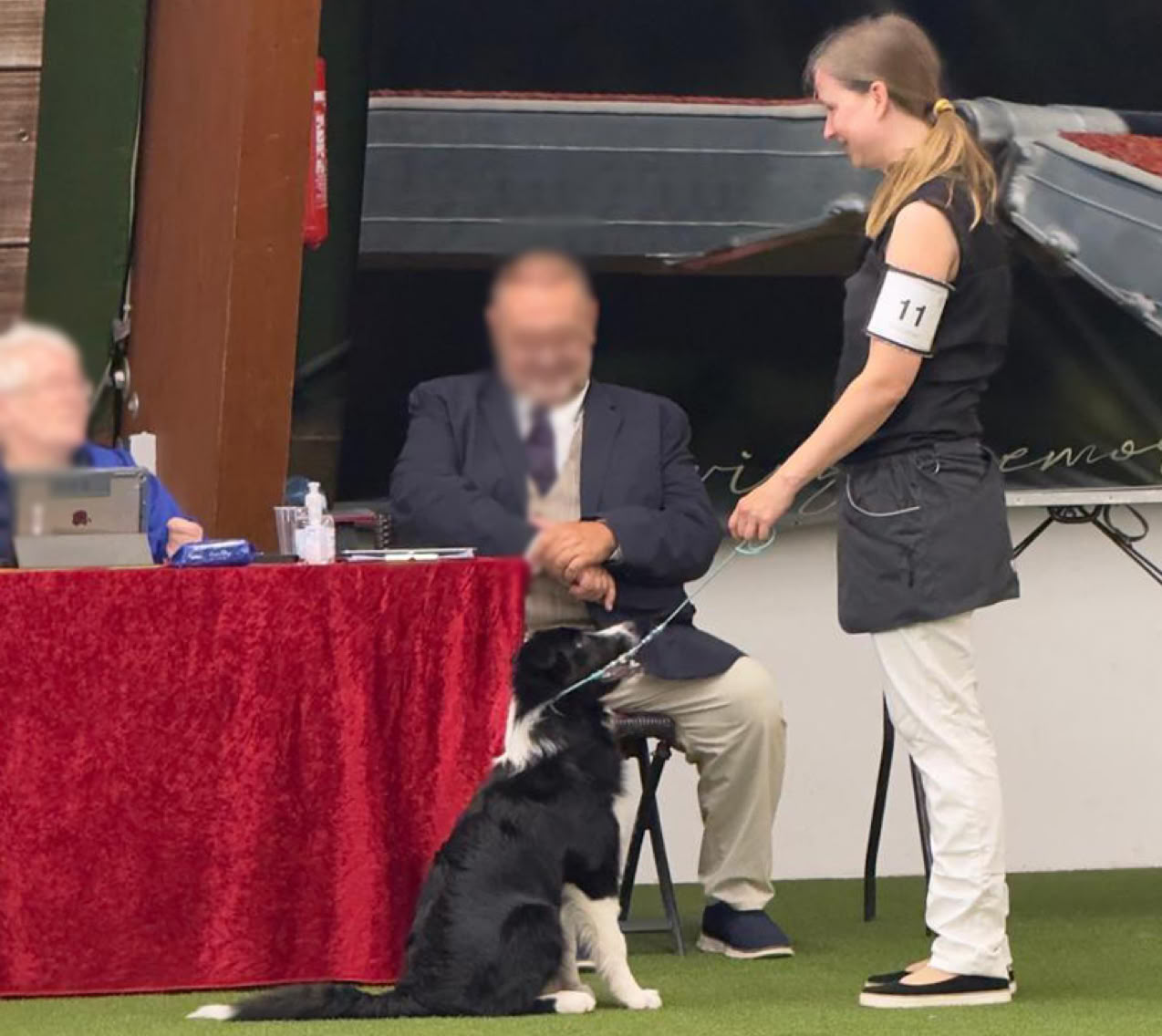 Eine Frau steht neben einem sitzenden Hund vor einem Tisch mit zwei sitzenden Personen, die hinter einem roten Tuch sitzen.