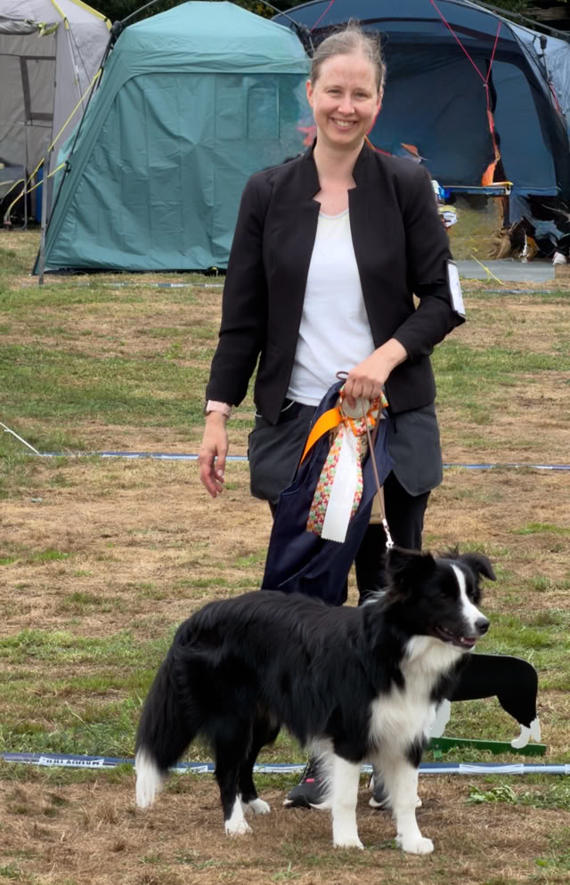 Eine Person steht auf einer Wiese neben einem schwarz-weißen Hund, im Hintergrund sind Zelte zu sehen.