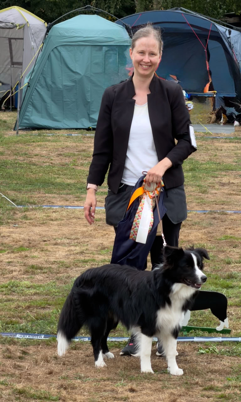 Eine Person steht auf einer Wiese neben einem schwarz-weißen Hund, im Hintergrund sind Zelte zu sehen.