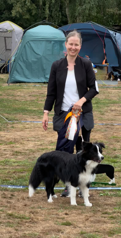 Eine Person steht auf einer Wiese neben einem schwarz-weißen Hund, im Hintergrund sind Zelte zu sehen.